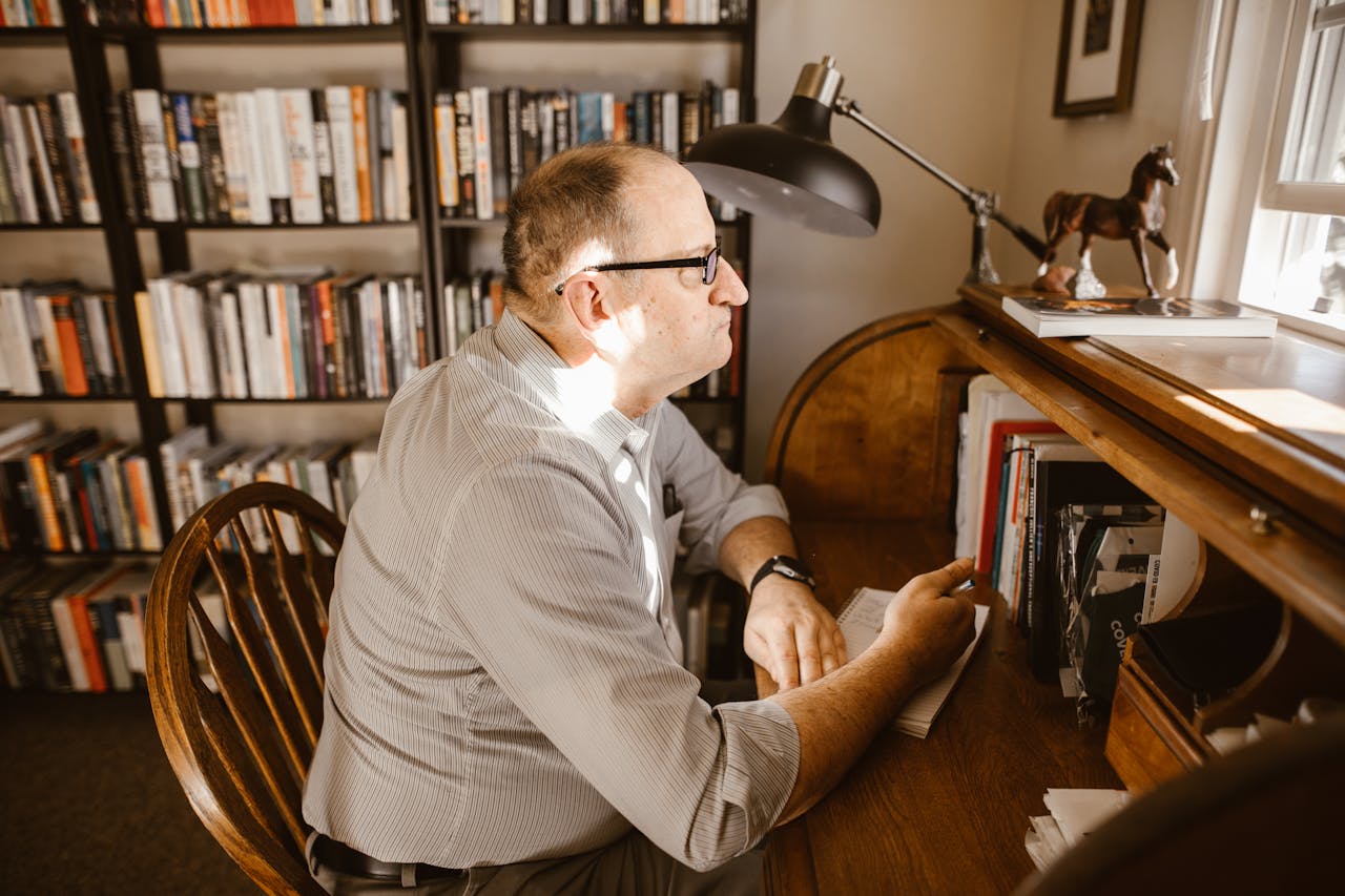 services-03 A man wearing glasses reads at his desk in a cozy home library filled with books.